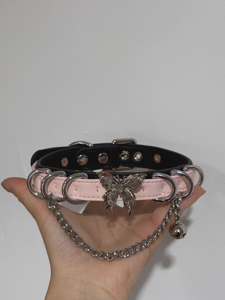 Black and pink leather choker featuring a stainless steel plated butterfly charm, D rings, chain and bell. Held by a hand against a white background for a kawaii aesthetic.