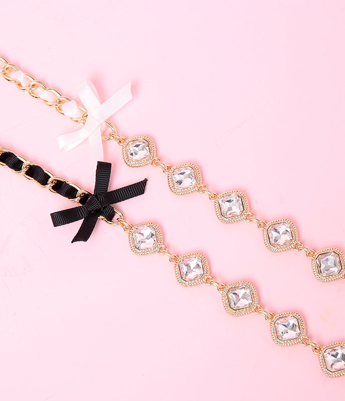 Close-up of two black and pink chain leashes for the Molly choker design, showing the black and pink ribbon bow appliques, ribbon threaded gold chain and clear crystal charms. Displayed on a pink background for a pet play aesthetic.
