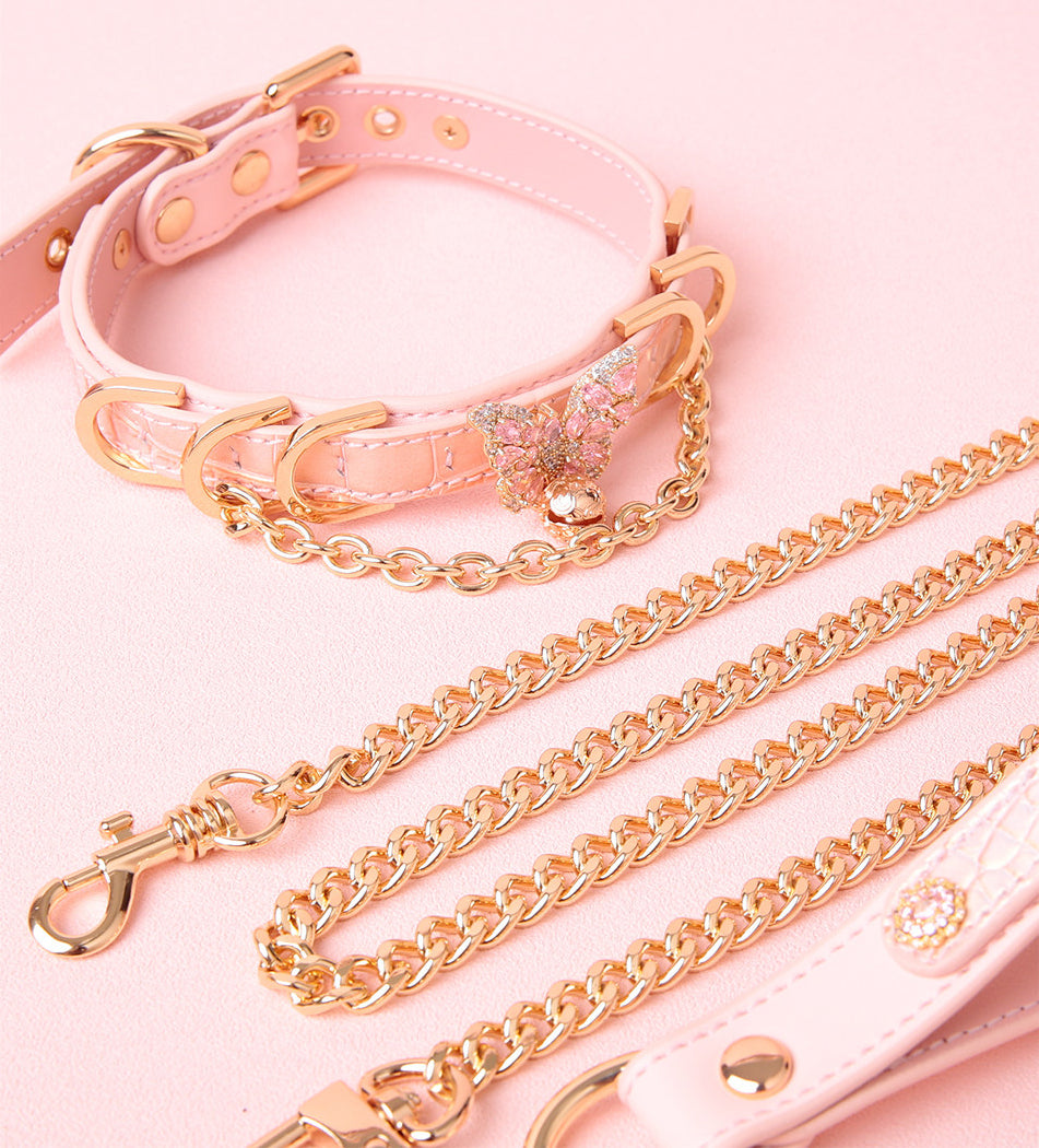 Pink leather choker and leash set featuring a gold and pink diamante butterfly charm, detachable gold chain leash and pink and gold leather hand strap. Displayed on a pink background for a pet play aesthetic.