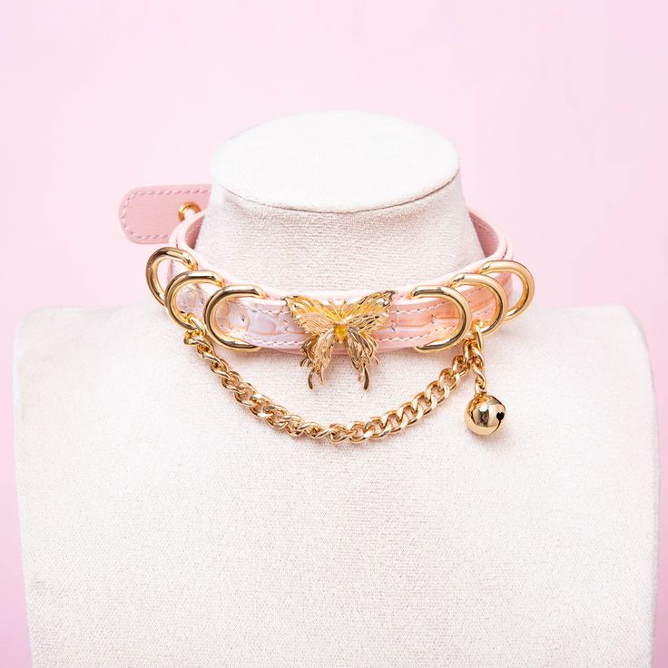 Pink leather choker featuring a gold tone butterfly charm, D rings, chain and bell. Displayed on a white mannequin bust against a pink background for a glam goth aesthetic.