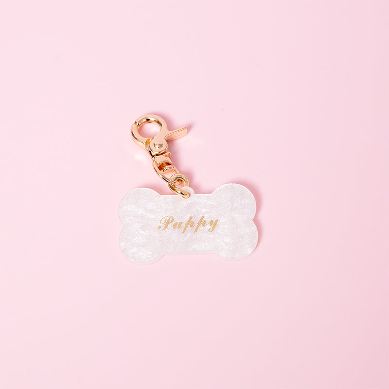 Detachable white acrylic bone shaped pet tag connected to a gold tone swivel lobster clasp, featuring gold foil engraving reading "Puppy" as an example of custom engraving available. Displayed on a light pink background for a kawaii aesthetic.