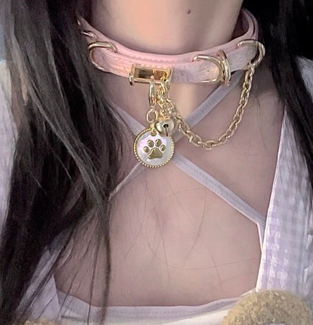 Worn photo of a pink leather choker featuring a white and gold paw print pet tag, gold tone chain, D rings and bell charm. Styled with brown pigtails and a pink and white gingham dress for a Harajuku fashion aesthetic.