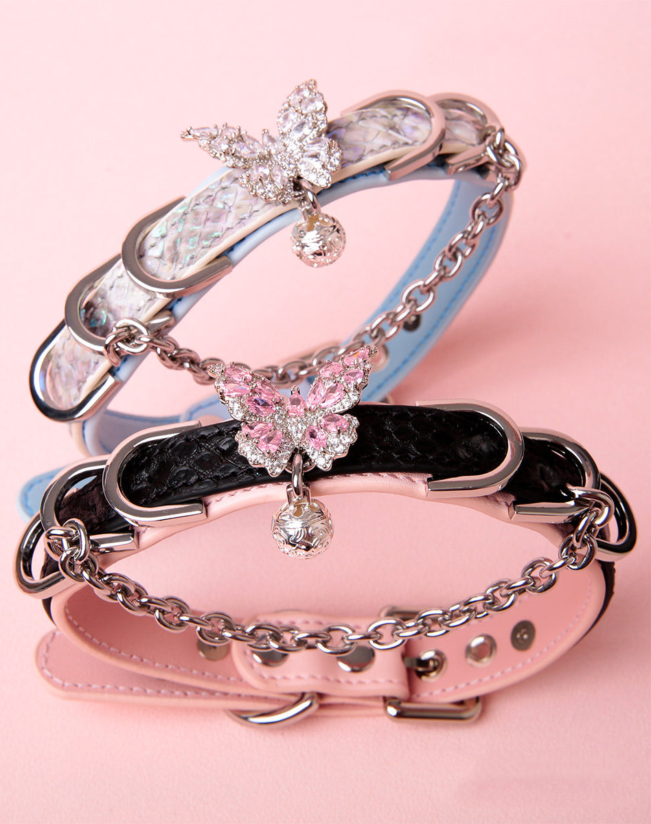 Two leather chokers, one black and pink, the other blue and silver, featuring silver diamante butterfly charms, chains, D rings and bells. Displayed on a pink background for a kawaii pet play aesthetic.