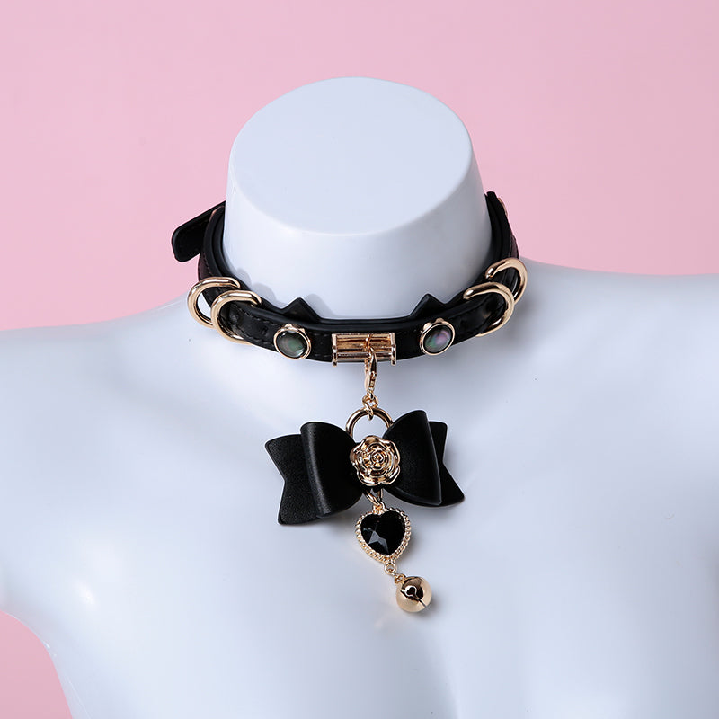 Black leather choker featuring a cat ear shaped strap, detachable leather bow charm with a black and gold crystal heart and bell charm. Displayed on a white mannequin against a pink background for an e-girl aesthetic.
