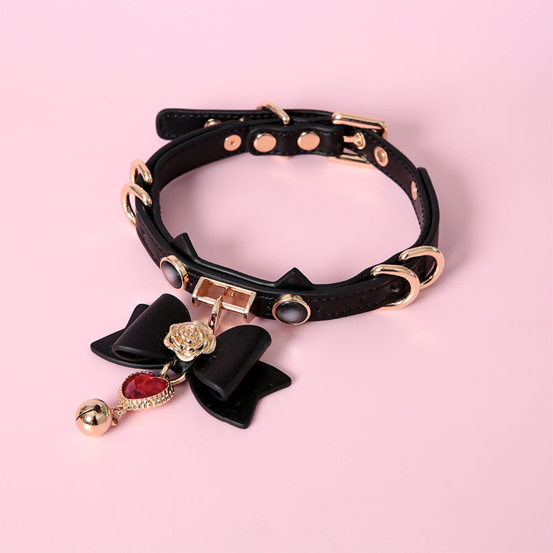 Black leather choker featuring a cat ear shaped strap, detachable leather bow pendant with a red and gold crystal heart and bell charm. Displayed against a pink background for a kawaii goth aesthetic.