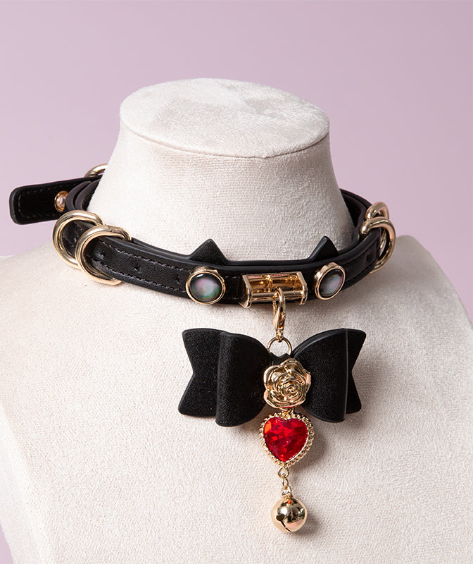 Black leather choker featuring a cat ear shaped strap, detachable leather bow charm with a red and gold crystal heart and bell charm. Displayed on a white mannequin bust against a pink background for a glam goth aesthetic.