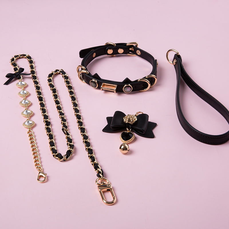 Black leather choker and leash set featuring cat ear and ribbon shaped elements, black and gold crystal heart and bell charm with detachable diamante, ribbon and chain leash and leather hand strap. Displayed against a pink background for a pastel goth aesthetic.
