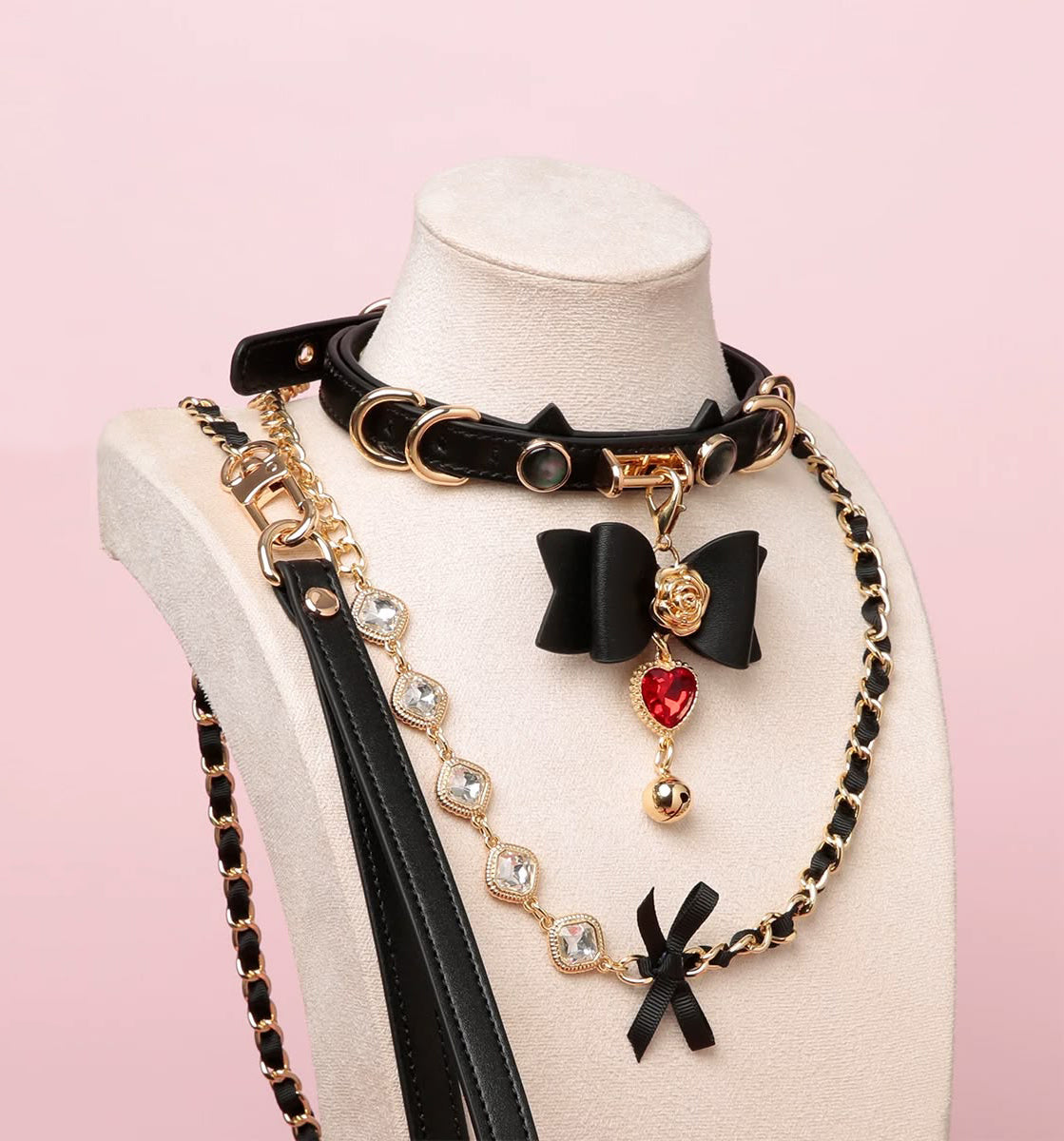 Black leather choker and leash set featuring cat ear and ribbon shaped elements, red and gold crystal heart and bell charm with detachable diamante, ribbon and chain leash and leather hand strap. Displayed on a white mannequin against a pink background for a kawaii goth aesthetic.