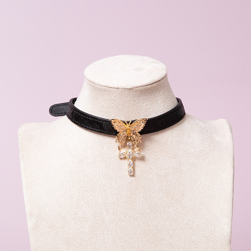 Black leather choker featuring an intricate gold tone butterfly pendant and gold and clear diamante cross charm. Displayed on a white mannequin bust against a pink background for a gothic gyaru aesthetic.