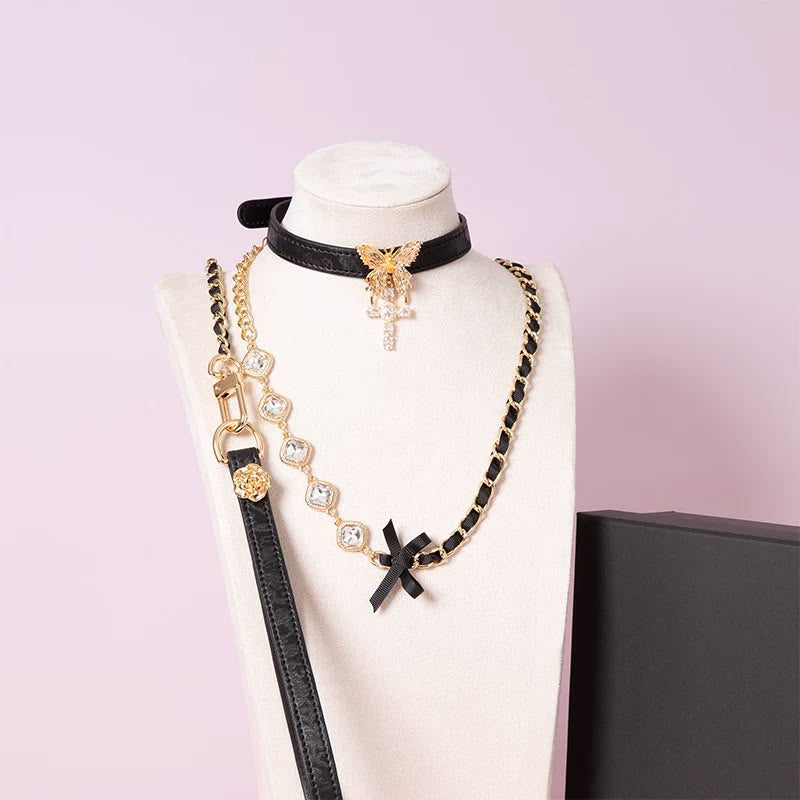 Black leather choker featuring an intricate gold tone butterfly pendant and gold and clear diamante cross charm, with detachable gold and crystal chain leash and black leather hand strap. Displayed on a white mannequin bust against a pink background for a glam goth aesthetic.