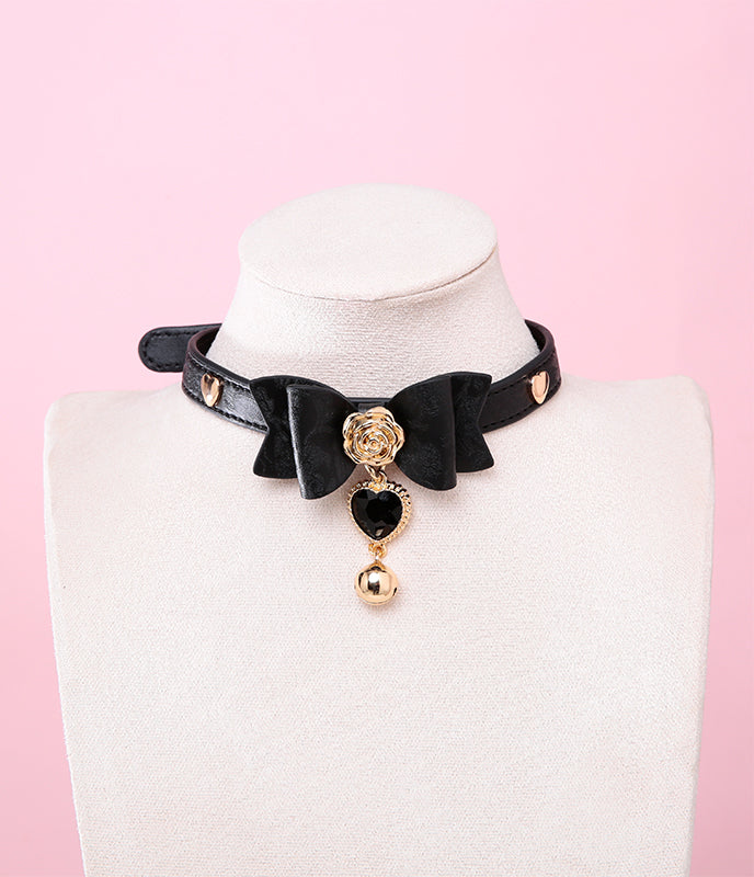 Black leather choker featuring a central bow shaped design, gold tone rose and heart studs with detachable black and gold crystal heart and bell charm. Displayed on a white mannequin bust against a pink background for a glam goth aesthetic.