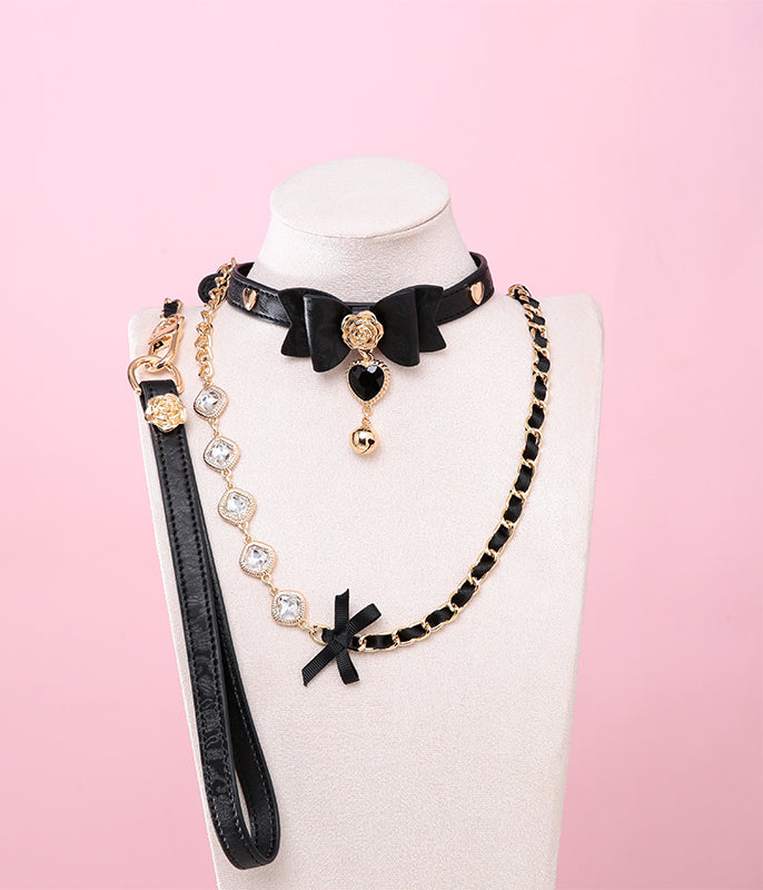 Black leather choker and leash set featuring a central bow shaped design, gold tone rose and heart studs, black and gold crystal heart and bell charm with detachable diamante, ribbon and chain leash and leather hand strap. Displayed on a white mannequin bust against a pink background for a kawaii goth aesthetic.
