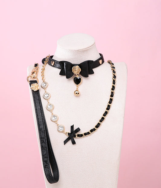 Black leather choker and leash set featuring a central bow shaped design, gold tone rose and heart studs, black and gold crystal heart and bell charm with detachable diamante, ribbon and chain leash and leather hand strap. Displayed on a white mannequin bust against a pink background for a kawaii goth aesthetic.