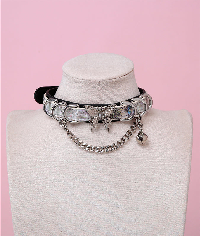 Black and holographic silver leather choker featuring a stainless steel plated butterfly charm, D rings, chain and bell. Displayed on a white mannequin bust against a pink background for a cybergoth aesthetic.