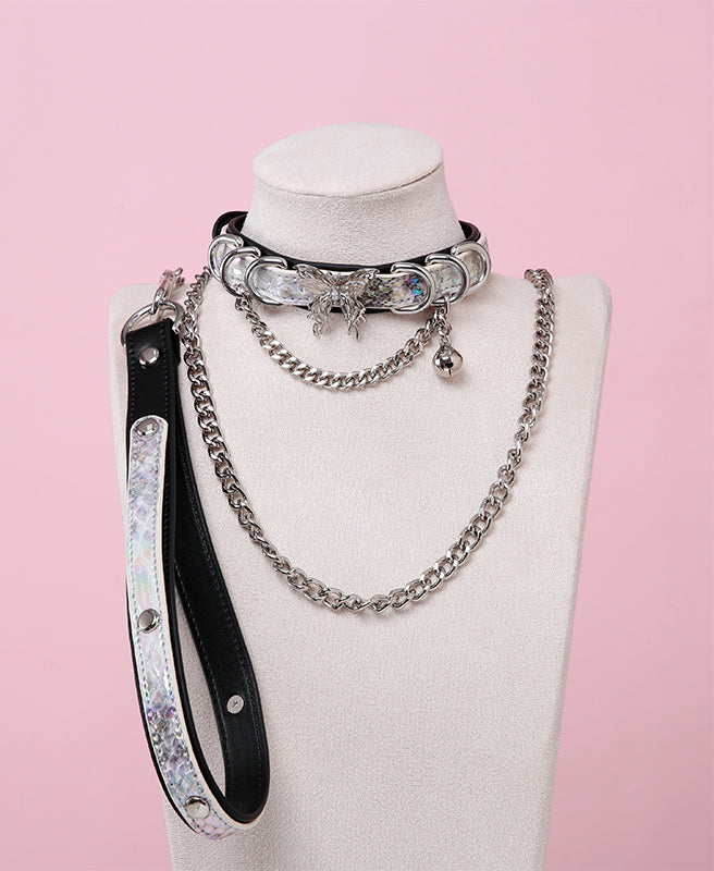 Black and holographic silver leather choker and leash set featuring stainless steel plated butterfly hardware, D rings, chain and bell with detachable chain leash and leather hand strap. Displayed on a white mannequin bust against a pink background for a Cybergoth aesthetic.