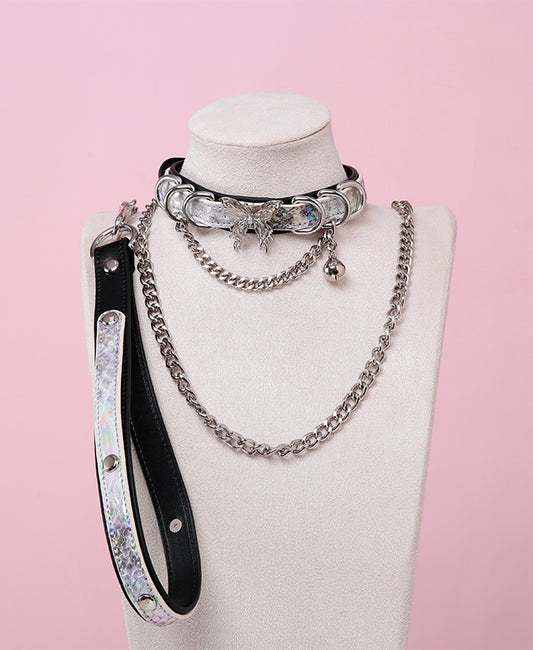Black and holographic silver leather choker and leash set featuring stainless steel plated butterfly hardware, D rings, chain and bell with detachable chain leash and leather hand strap. Displayed on a white mannequin bust against a pink background for a Cybergoth aesthetic.