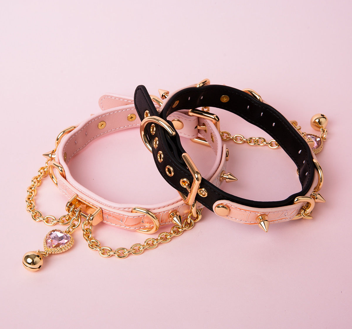 Two leather chokers, one black and pink, the other pink, featuring gold tone spike studs, chains, D rings and pink and gold crystal heart and bell charms. Displayed on a pink background for a kawaii pet play aesthetic.