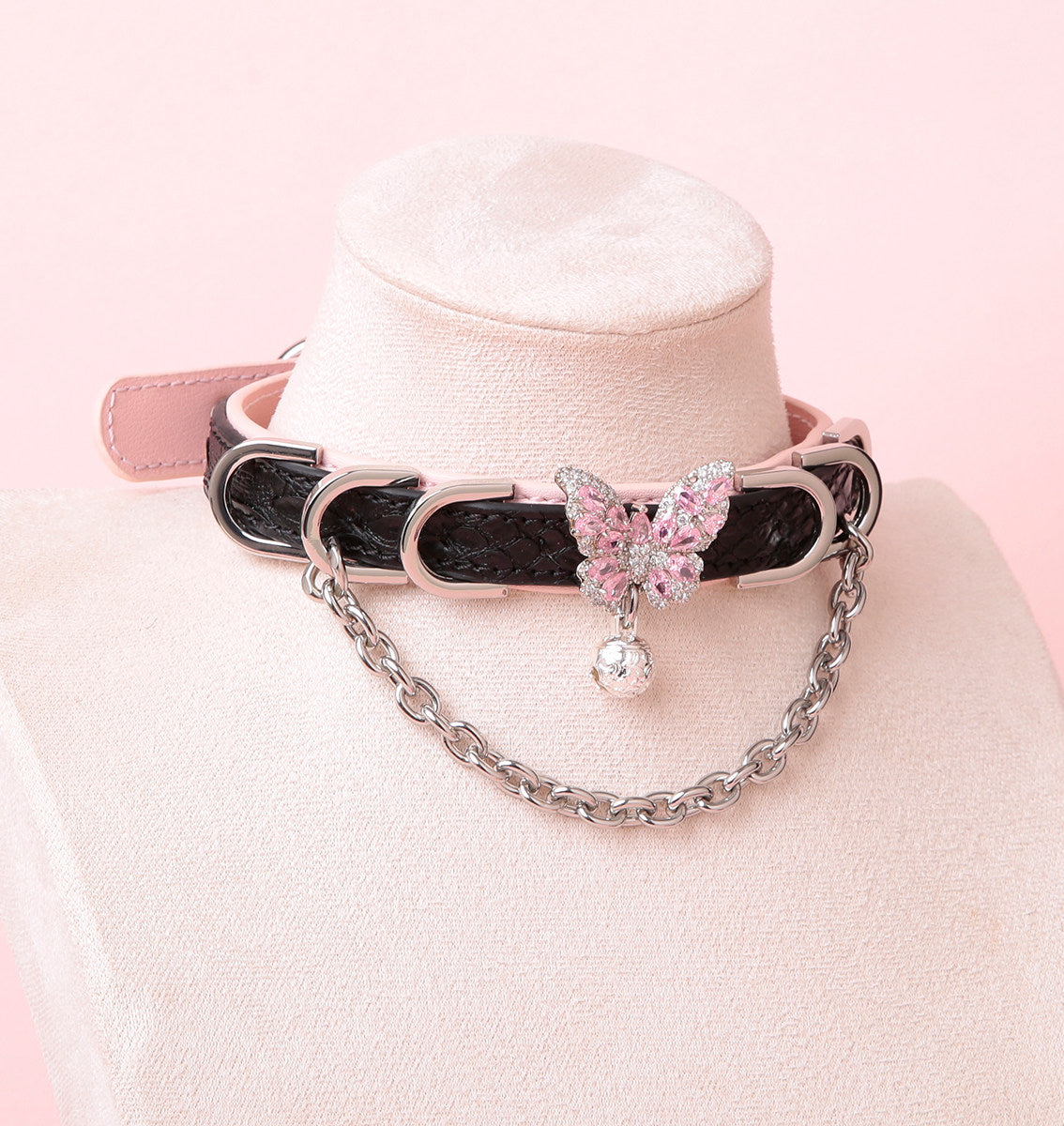 Black and pink leather choker featuring a silver and pink diamante butterfly charm, silver tone chains, D rings and bell. Displayed on a white mannequin bust against a pink background for a Harajuku fashion aesthetic.