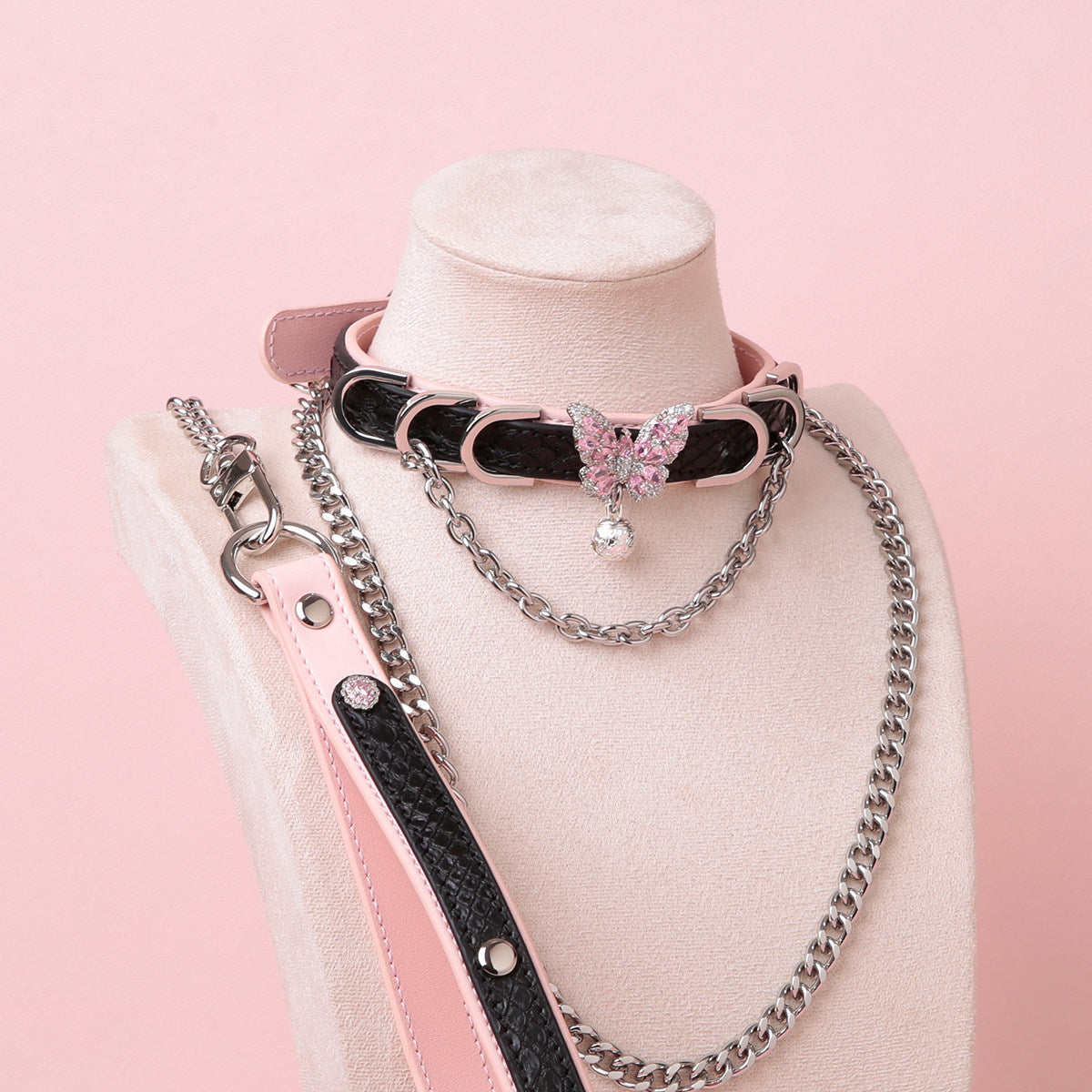 Black and pink leather choker and leash set featuring a silver and pink diamante butterfly charm, silver chains, D rings and bell, with detachable chain leash and leather hand strap. Displayed on a white mannequin bust against a pink background for a pastel goth aesthetic.