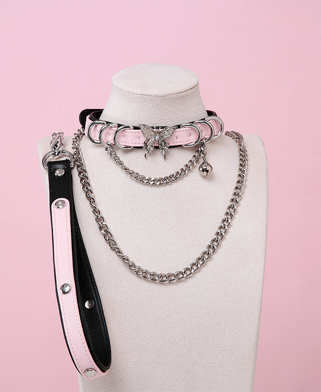 Black and pink leather choker and leash set featuring stainless steel plated butterfly hardware, D rings, chain and bell with detachable chain leash and leather hand strap. Displayed on a white mannequin bust against a pink background for a pastel goth aesthetic.