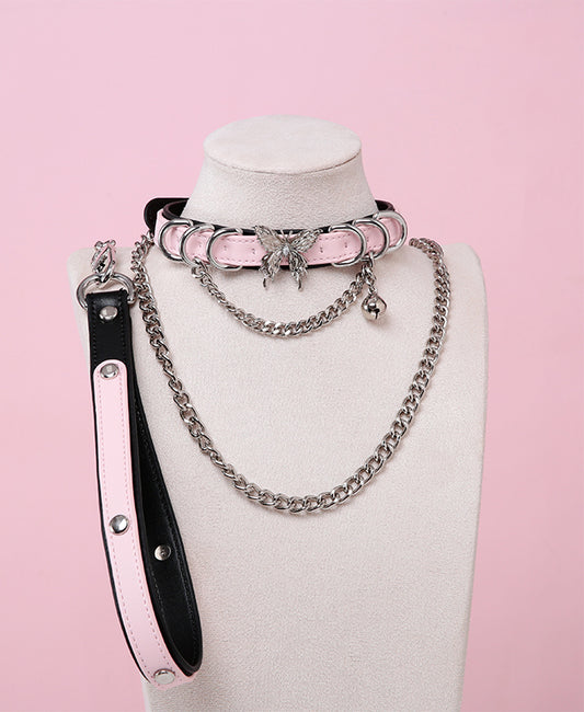 Black and pink leather choker and leash set featuring stainless steel plated butterfly hardware, D rings, chain and bell with detachable chain leash and leather hand strap. Displayed on a white mannequin bust against a pink background for a pastel goth aesthetic.