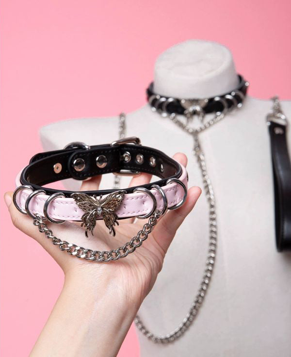 Black and pink leather choker featuring a stainless steel plated butterfly charm, D rings, chain and bell, with a detachable chain leash and leather hand strap. Held by a hand against a white mannequin bust and pink background for a kawaii aesthetic.