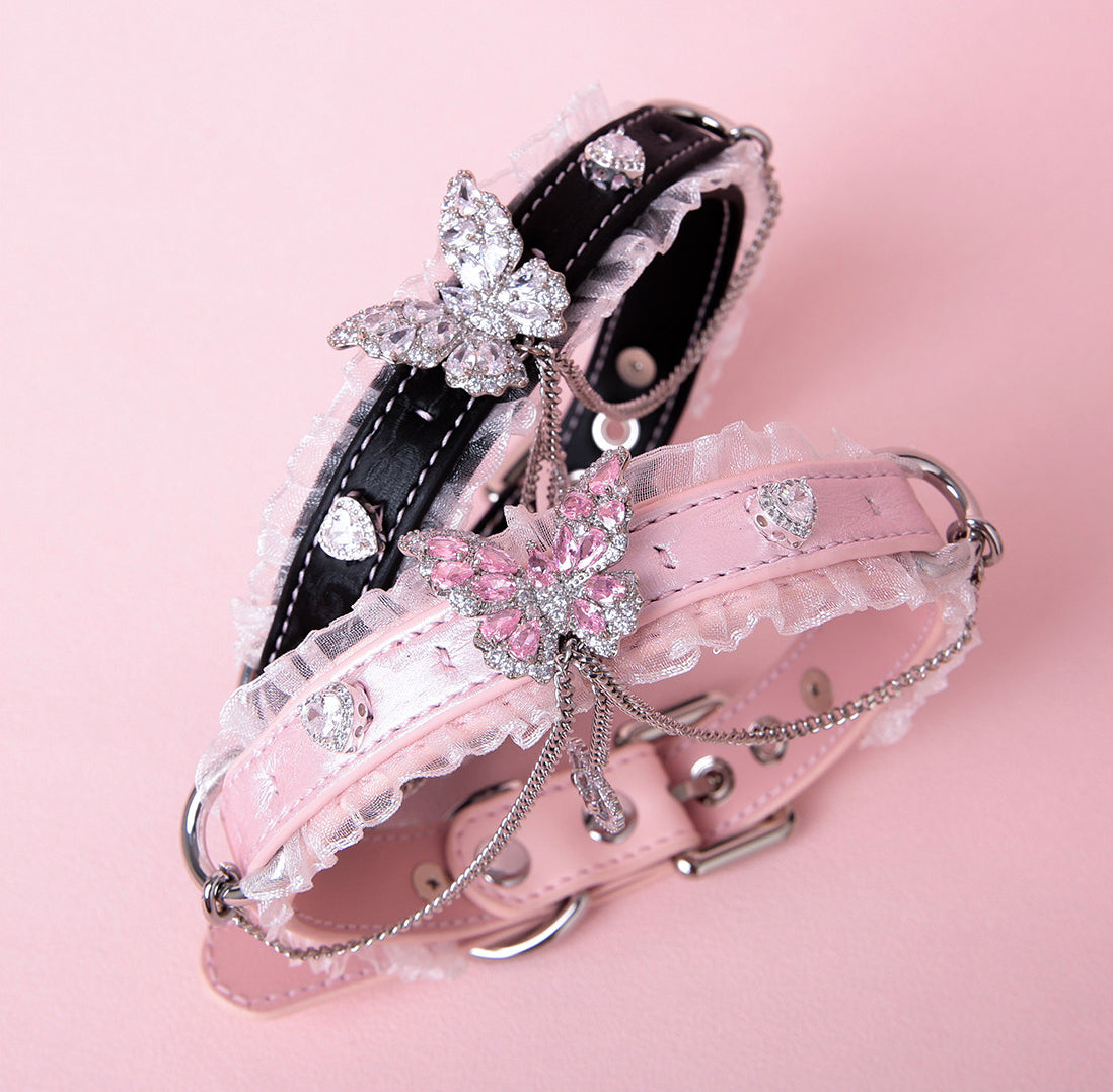 Two black and pink leather chokers featuring white ruffled lace trim, diamante butterfly and heart charms, silver tone chain and D rings. Displayed against a pink background for a Harajuku fashion aesthetic.