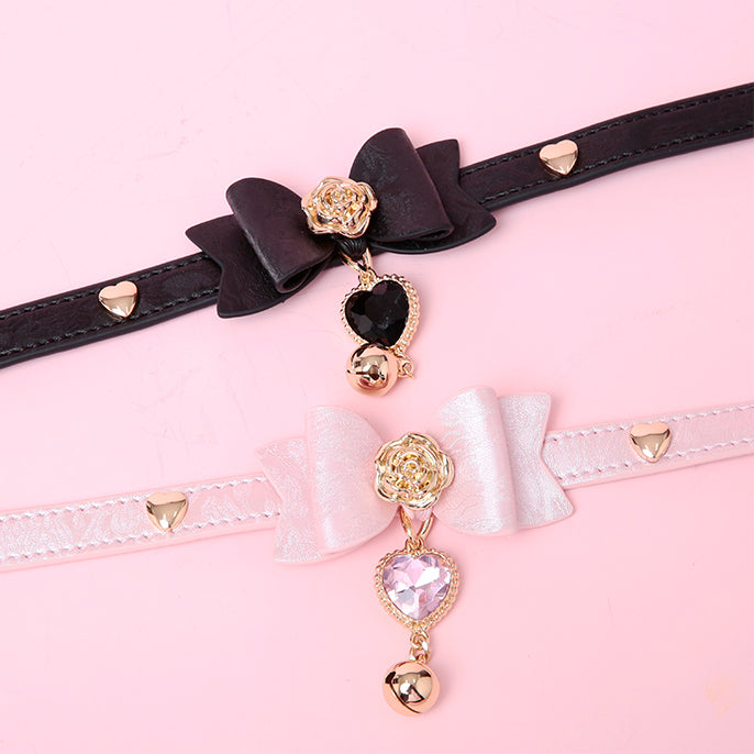 Close-up of two black and pink leather chokers featuring a central bow shaped design, gold tone rose and heart studs with detachable crystal heart and bell charms. Displayed on a white mannequin bust against a pink background for a kawaii goth aesthetic.