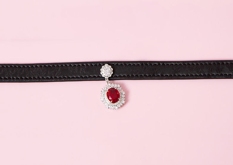 Close-up of a black leather choker showing the silver and red crystal cameo and hardware. Displayed on a pink background for a kawaii fashion aesthetic.