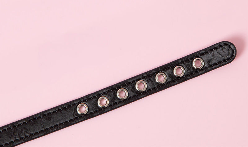 Close-up of a black leather choker showing multiple stainless steel plated eyelet holes, ensuring a durable and versatile fit. Displayed on a pink background for a kawaii fashion aesthetic.