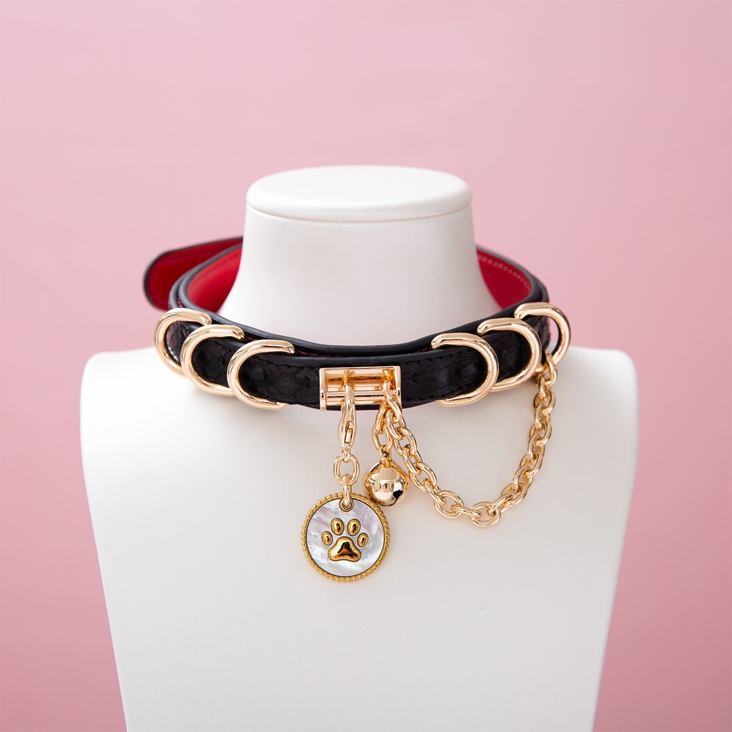 Black and red leather choker featuring a gold and white paw print tag, gold tone D rings, chain and bell. Displayed on a white mannequin bust against a pink background for a Harajuku fashion aesthetic.