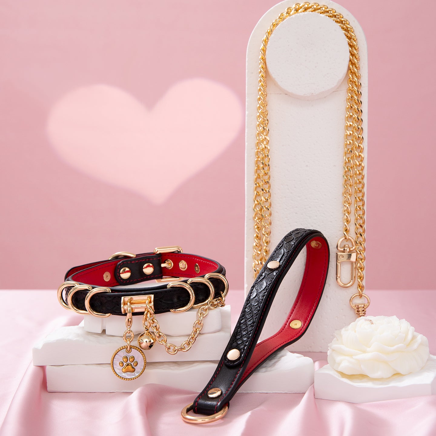 Black and red leather choker and leash set featuring a white and gold paw print tag, D rings, chain and bell with detachable chain leash and leather hand strap. Displayed on a white display stand against a pink background for a glam goth aesthetic.
