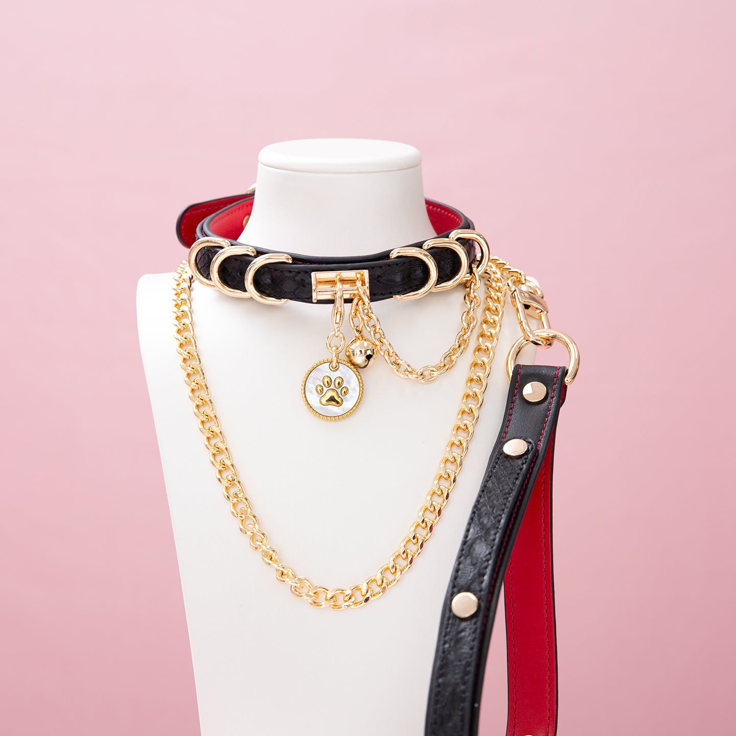 Black and red leather choker and leash set featuring a white and gold paw print tag, D rings, chain and bell with detachable chain leash and leather hand strap. Displayed on a white mannequin bust against a pink background for a goth gyaru aesthetic.