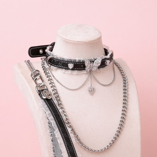 Black leather choker featuring white ruffled lace trim, diamante butterfly and heart charms, silver tone chain and D rings with detachable silver chain leash and black and white leather hand strap. Displayed on a white mannequin bust against a pink background for a pastel goth aesthetic.
