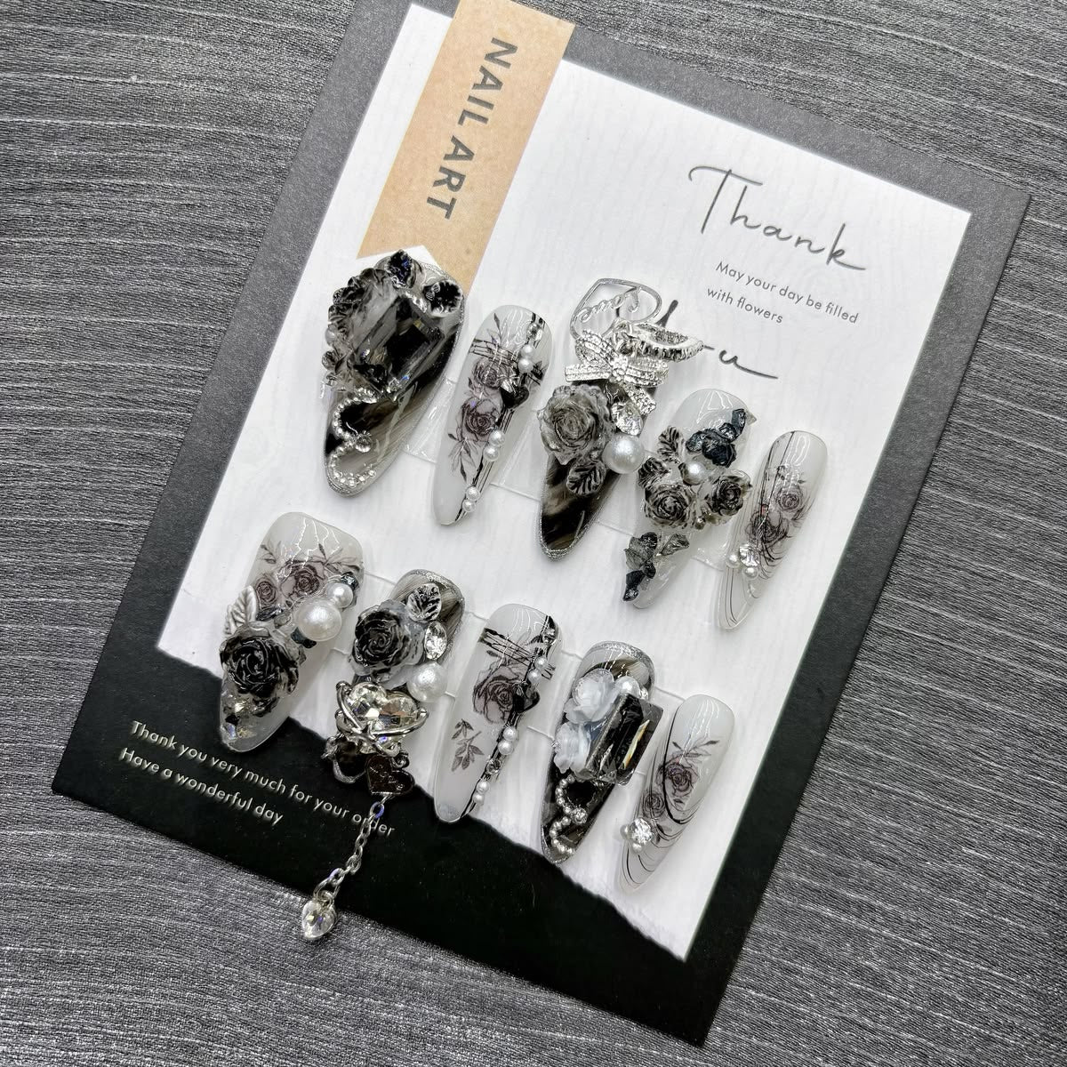 A set of decorative press-on nails displayed on a printed card, photographed on a gray wood-textured surface. The nails feature black, white, and gray floral and abstract designs with clear resin sections and small metallic details.