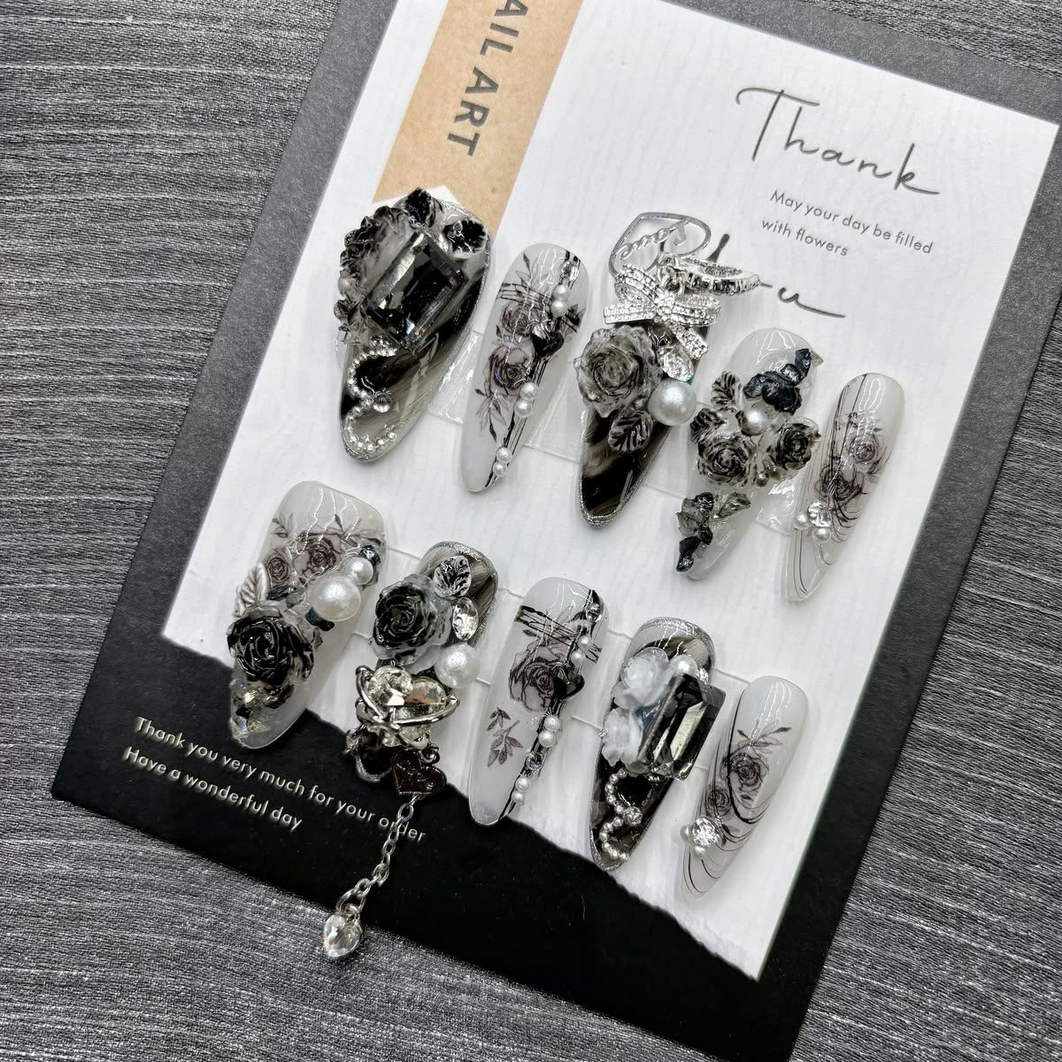 Black and white long stiletto false nails featuring a gothic rose pattern, silver and diamante bow and heart charms, white pearl beads and clear diamantes, displayed on a black and white display card.
