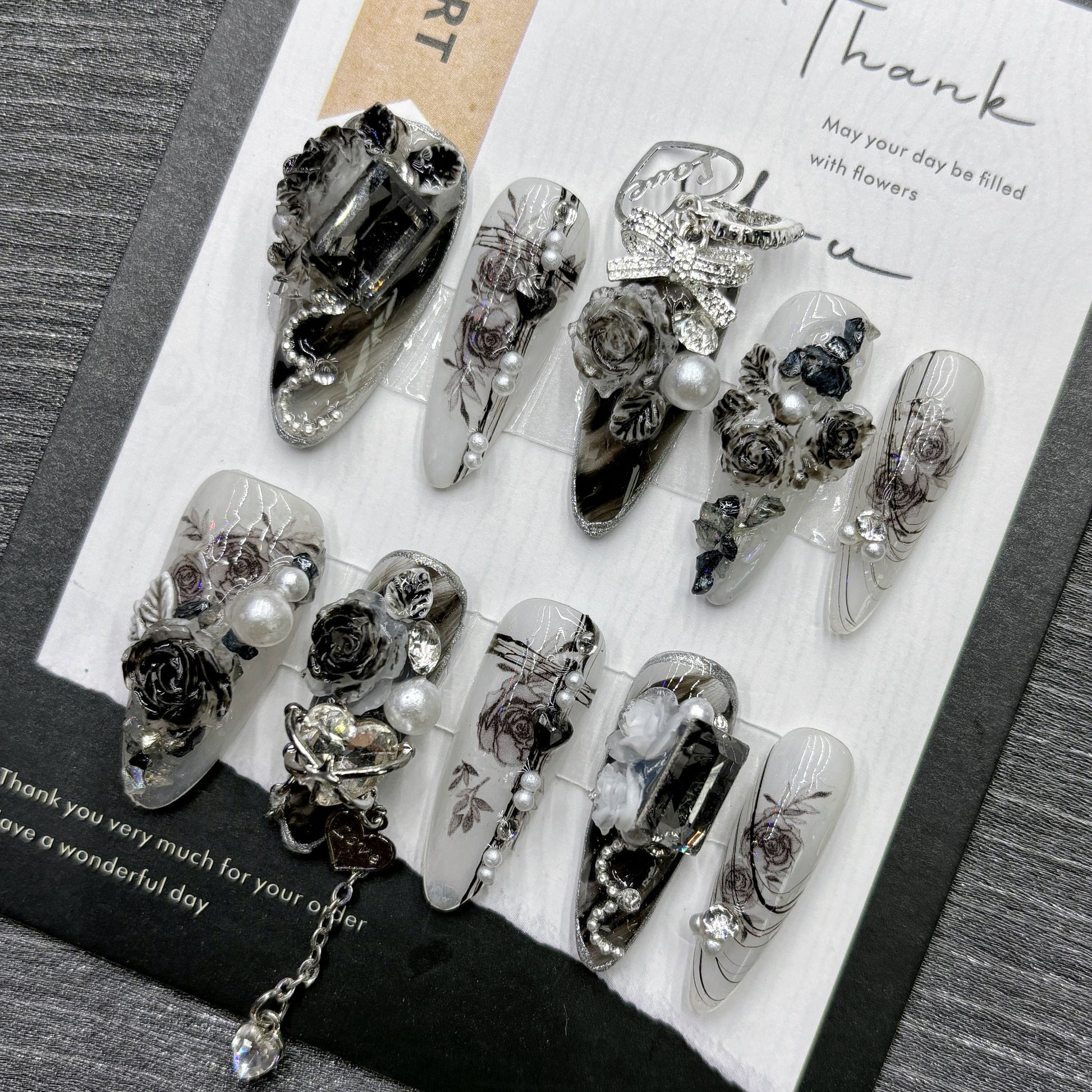 Black and white long stiletto false nails featuring a gothic rose pattern, silver and diamante bow and heart charms, white pearl beads and clear diamantes, displayed on a black and white display card.