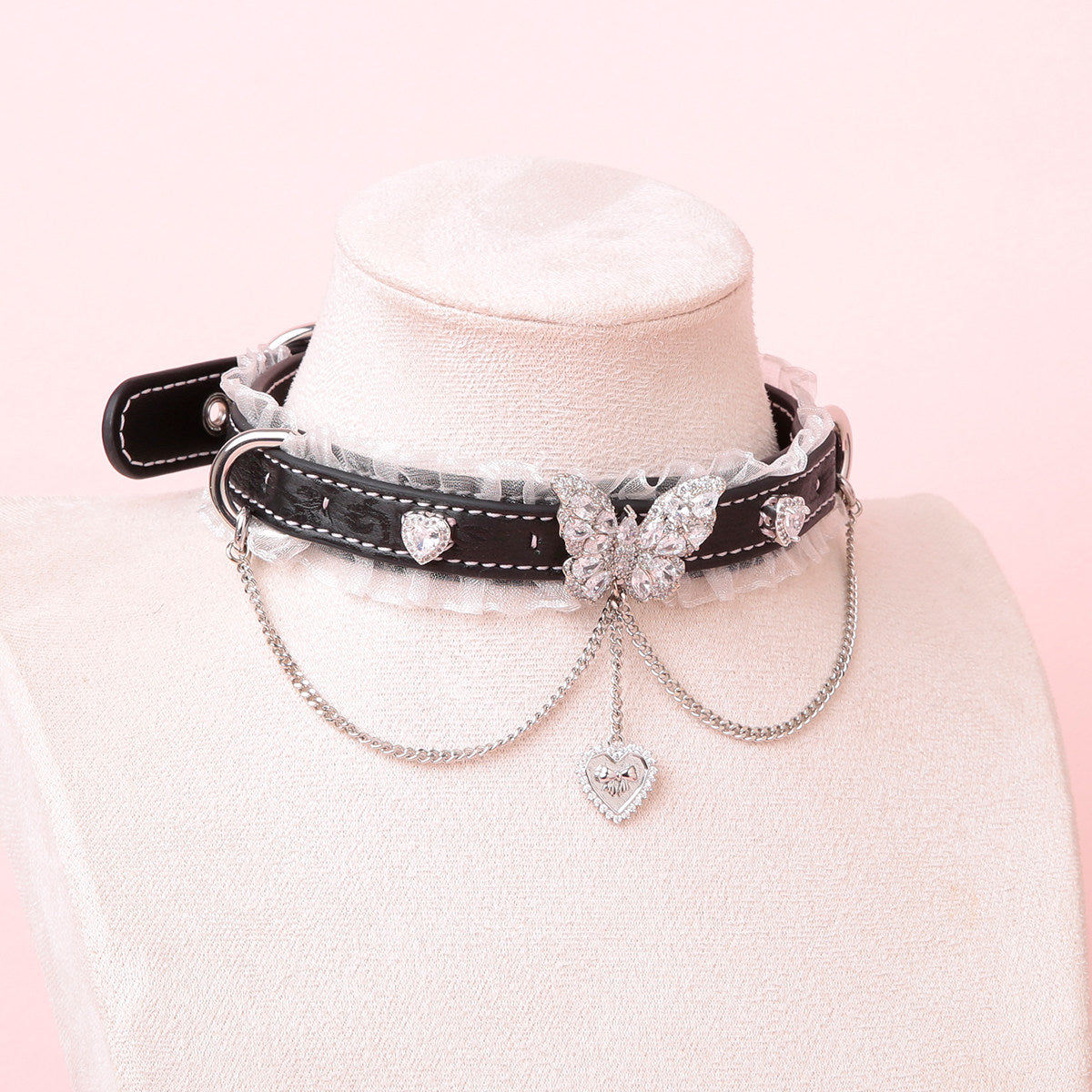 Black leather choker featuring white ruffled lace trim, diamante butterfly and heart studs, silver tone D rings and chain. Displayed on a white mannequin bust against a pink background for a kawaii goth aesthetic.
