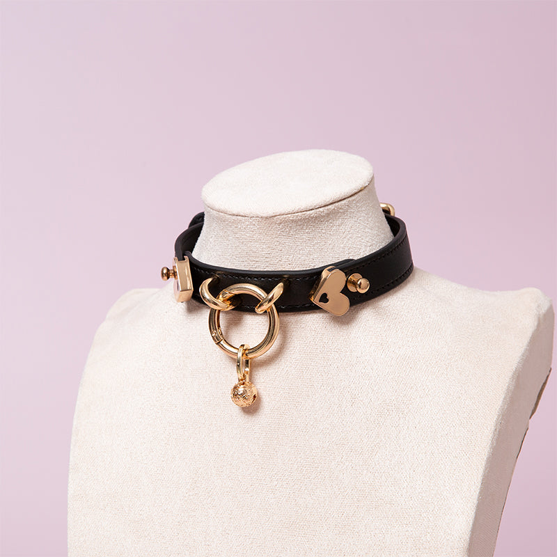 Black leather choker featuring gold tone O ring and heart shaped hardware and detachable ornate bell charm. Displayed on a white mannequin bust against a light pink background for a pastel goth aesthetic.