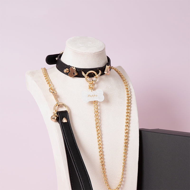 Black leather choker featuring gold tone hardware, custom engraved bone shaped pet tag, heavy duty chain leash and leather hand strap. Displayed on a white mannequin bust against a light pink background for a pastel goth aesthetic.