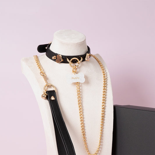 Black leather choker featuring gold tone hardware, custom engraved bone shaped pet tag, heavy duty chain leash and leather hand strap. Displayed on a white mannequin bust against a light pink background for a pastel goth aesthetic.