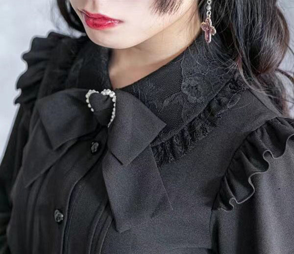 Close-up worn photo of a black long sleeved blouse, showing the lace trimmed Peter Pan style collar, ruffled shoulders, heart shaped buttons, pintucks and detachable bow brooch. Styled with black curly pigtails and red glossy lips for a kawaii aesthetic.