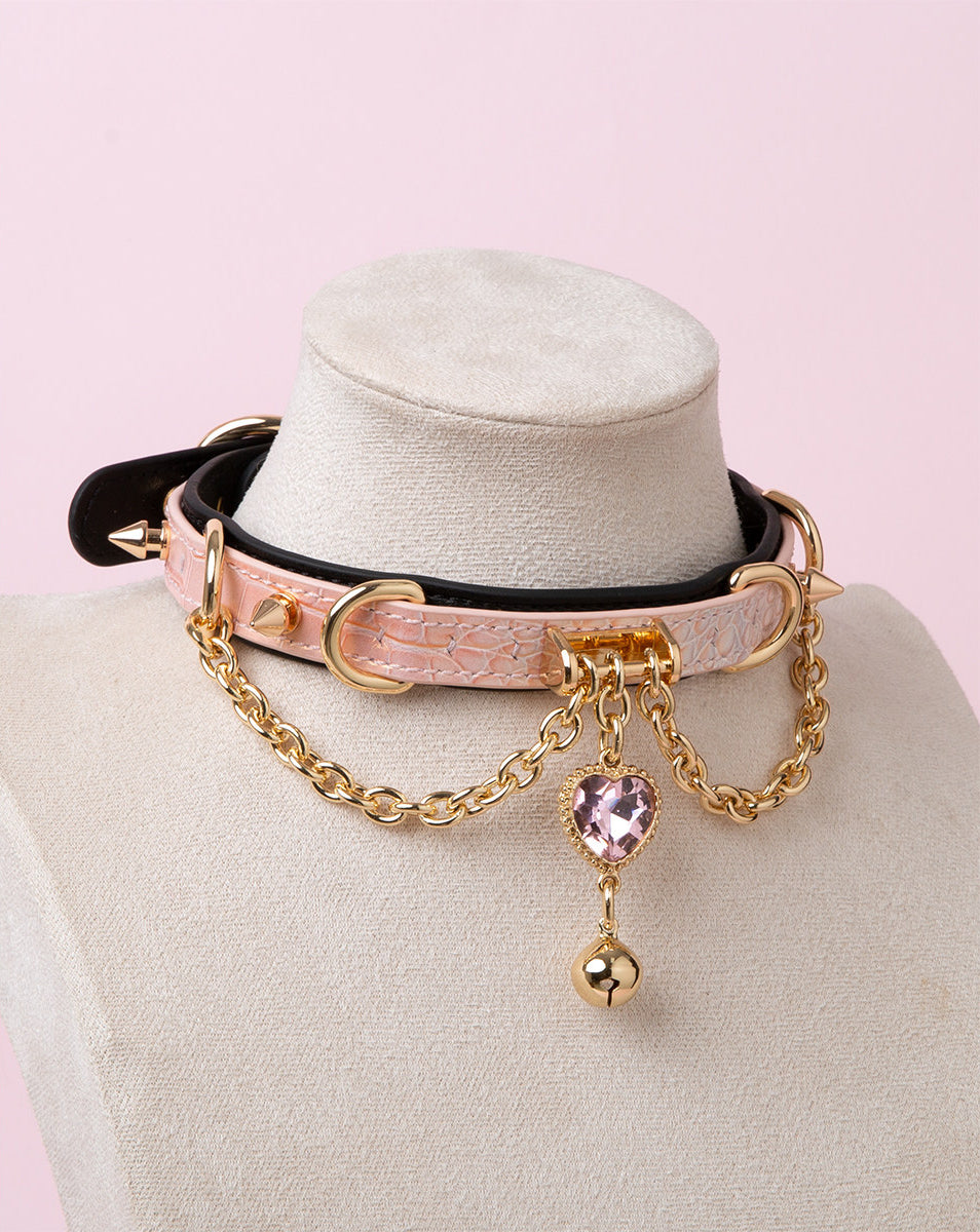 Black and pink leather choker featuring gold tone chains, D rings, spike studs, bell and pink and gold crystal heart charm. Displayed on a white mannequin bust against a pink background for a Harajuku fashion aesthetic.