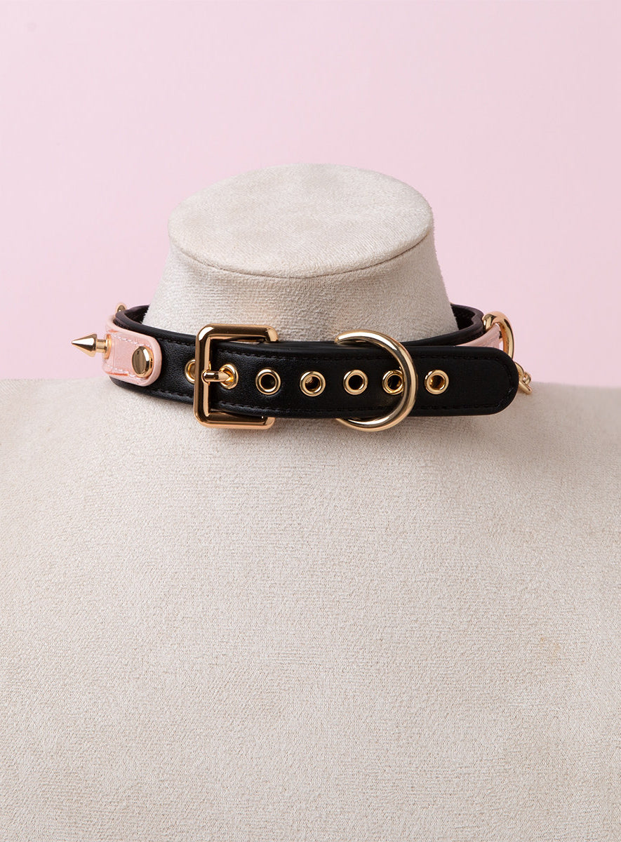 Rear view of a black and pink leather choker showing the adjustable gold tone buckle and eyelet holes, rivets and spike studs. Displayed on a white mannequin bust against a pink background for a Harajuku fashion aesthetic.