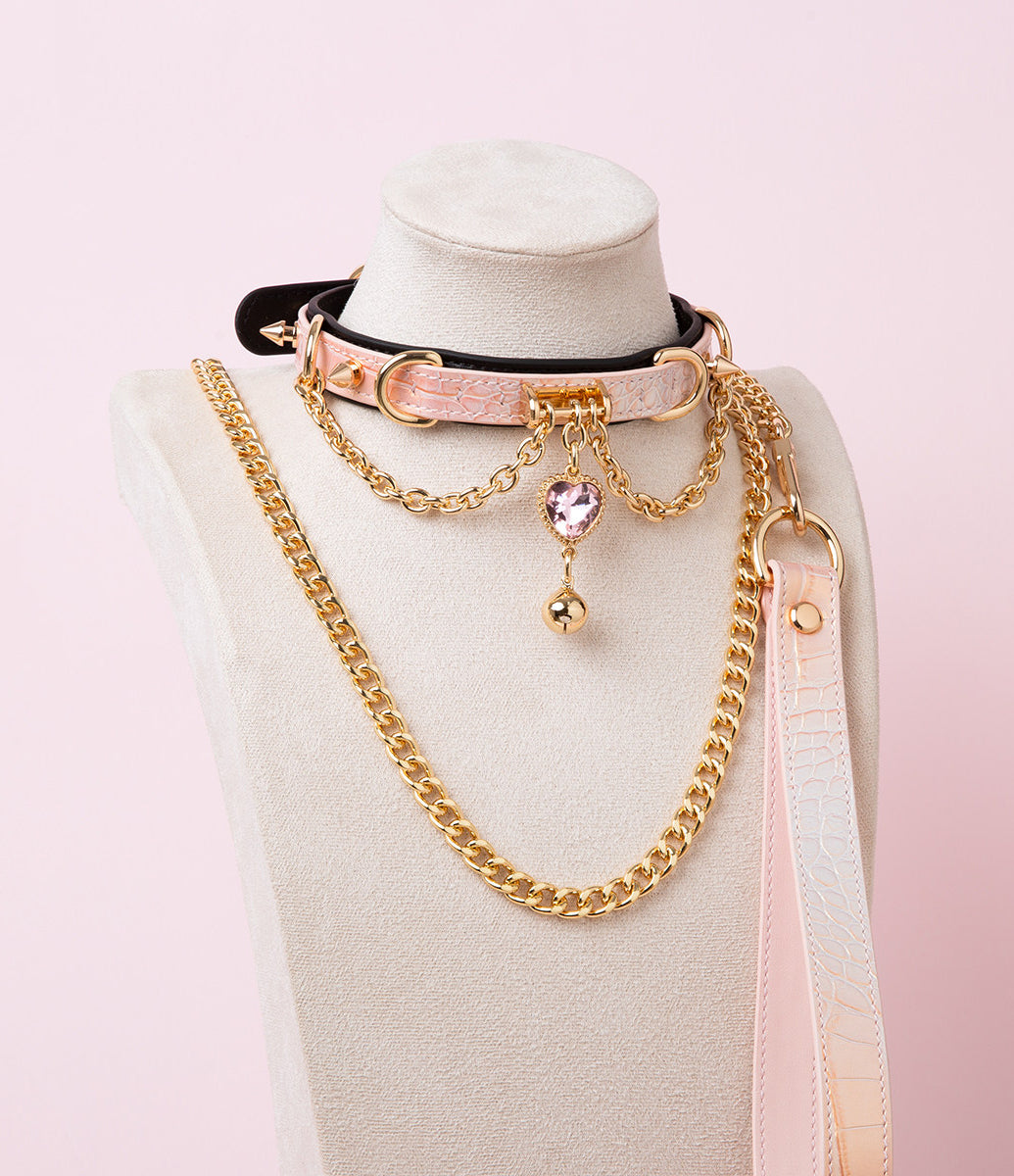 Black and pink leather choker and leash set featuring gold tone chains, D rings, spike studs, bell and pink and gold crystal heart charm, with detachable chain leash and leather hand strap. Displayed on a white mannequin bust against a pink background for a pastel goth aesthetic.