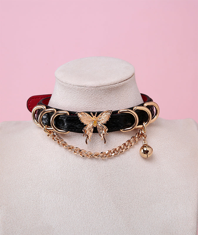 Black and red leather choker featuring a gold tone butterfly charm, D rings, chain and bell. Displayed on a white mannequin bust against a pink background for a glam goth aesthetic.