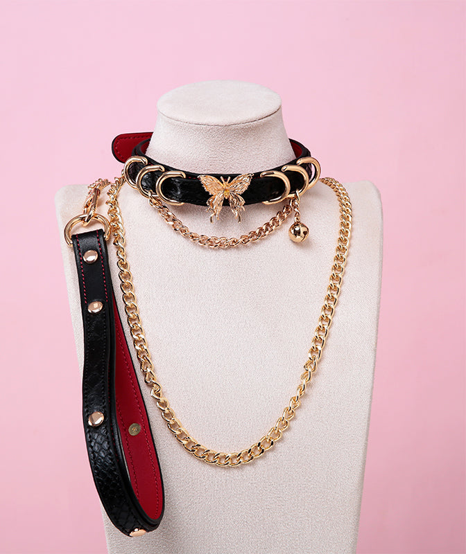 Black and red leather choker and leash set featuring gold tone butterfly charm, D rings, chain and bell with detachable chain leash and leather hand strap. Displayed on a white mannequin bust against a pink background for a gothic gyaru aesthetic.
