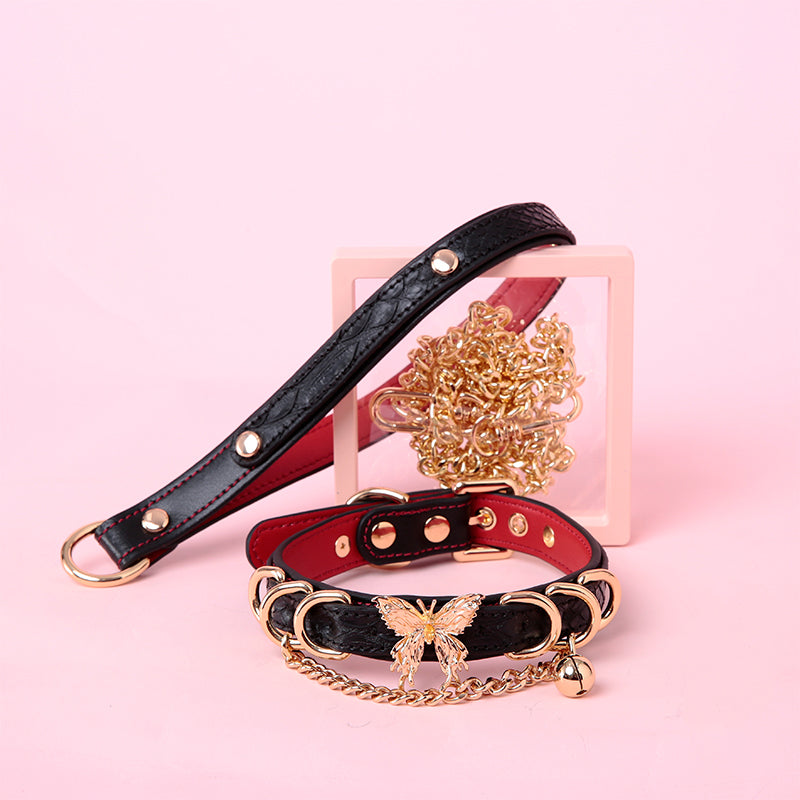 Black and red leather choker and leash set featuring an ornate gold tone butterfly charm, D rings, chain and bell charm with detachable chain leash and leather hand strap. Displayed against a pink background for a glam goth aesthetic.