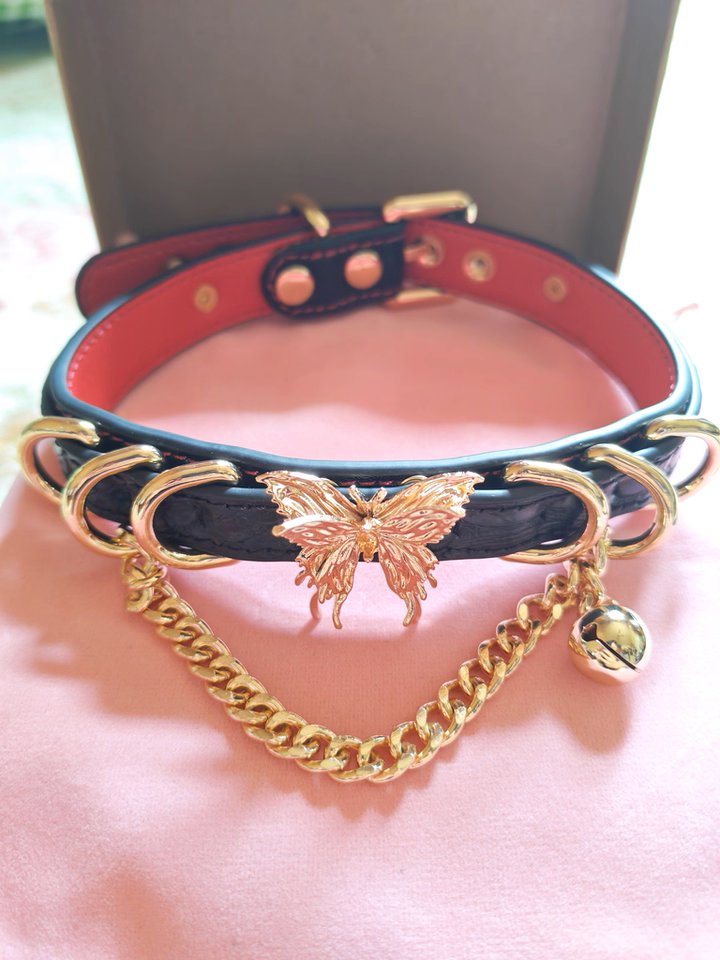 Black and red leather choker featuring a gold tone butterfly charm, D rings, chain and bell charm. Displayed on a pink velvet drawstring bag to show the packaging in a lifestyle shot.