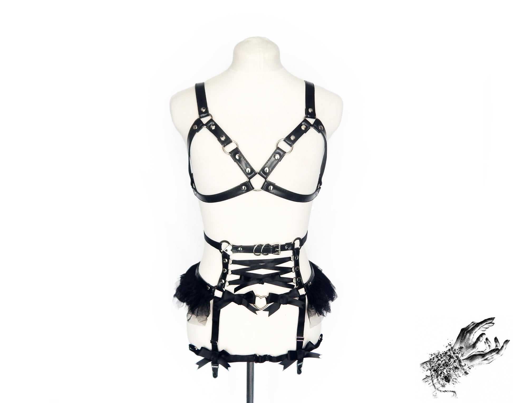 Front view of a black faux leather harness set featuring a separate harness bra and garter belt with silver O ring hardware, corset lacing and ruffled tulle displayed on a mannequin against a white background.